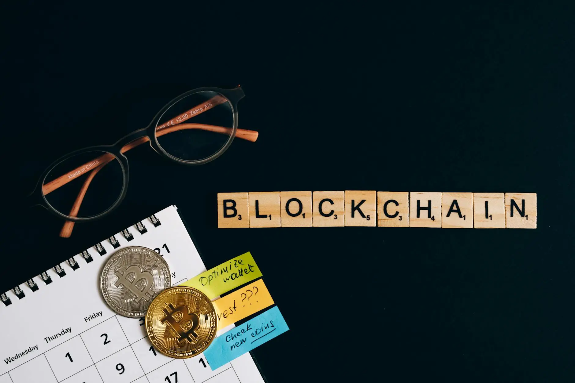 Wooden blocks spelling 'BLOCKCHAIN' with glasses and Bitcoin coins on a calendar.