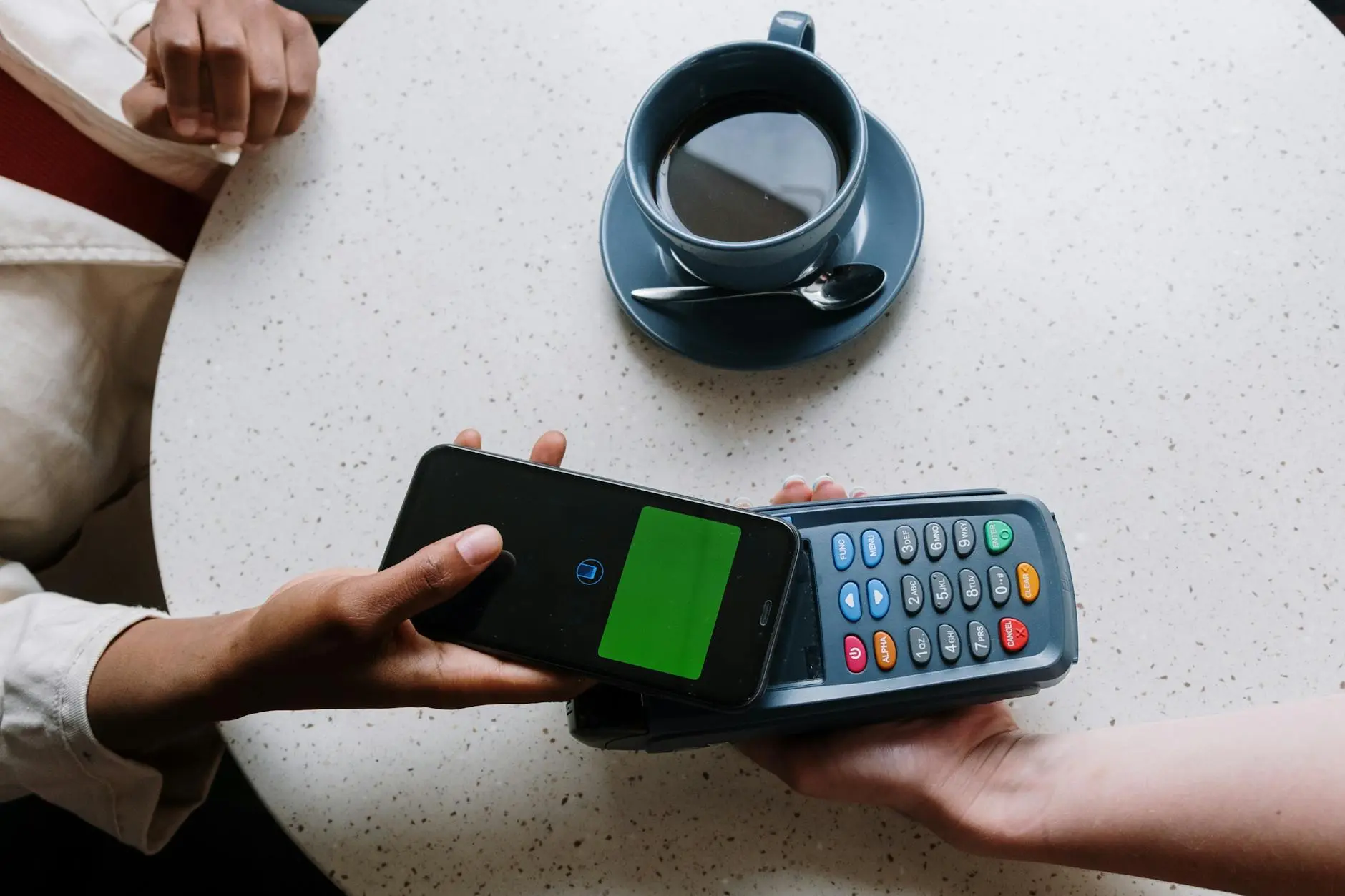 A smartphone with payment screen next to a card reader on a table with a cup of coffee.