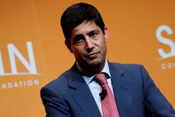 Portrait of Kevin Warsh wearing a blue suit and red patterned tie against an orange background.