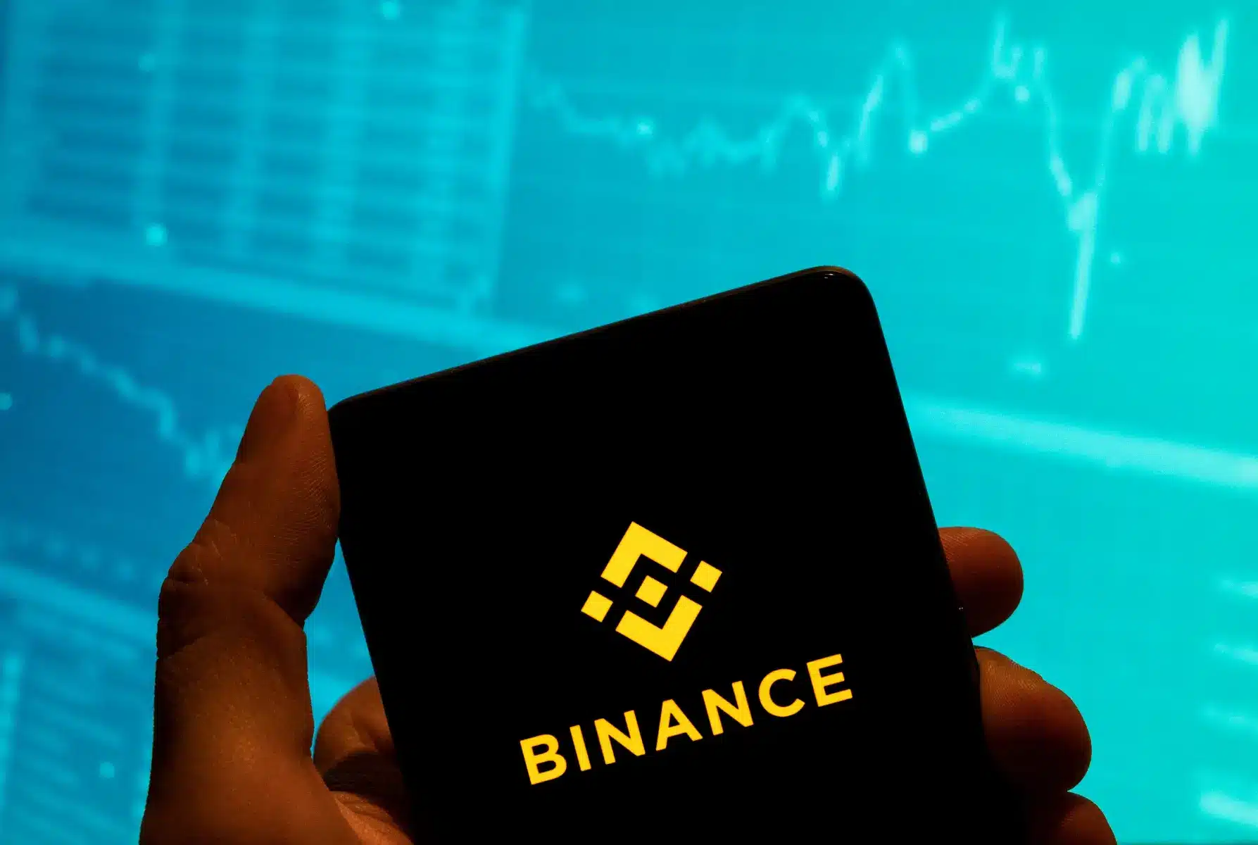 A hand holding a smartphone displaying the Binance logo and name against a backdrop of blue trading charts