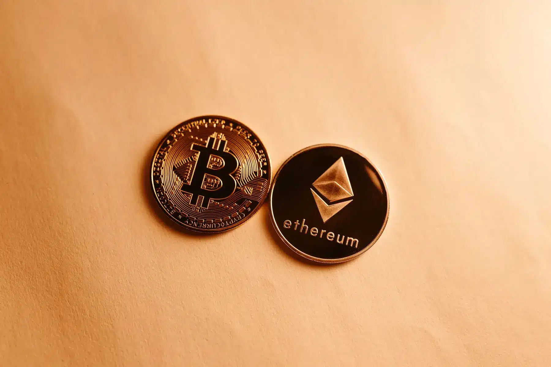 Physical gold Bitcoin and silver Ethereum coins resting on a textured light orange surface