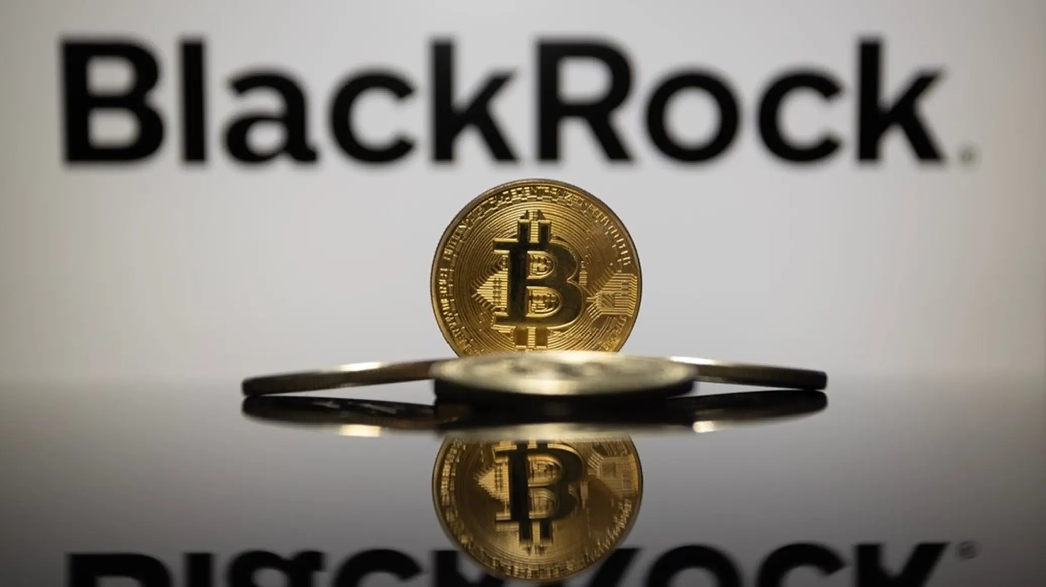 A physical gold Bitcoin coin standing upright in front of a blurred BlackRock company logo