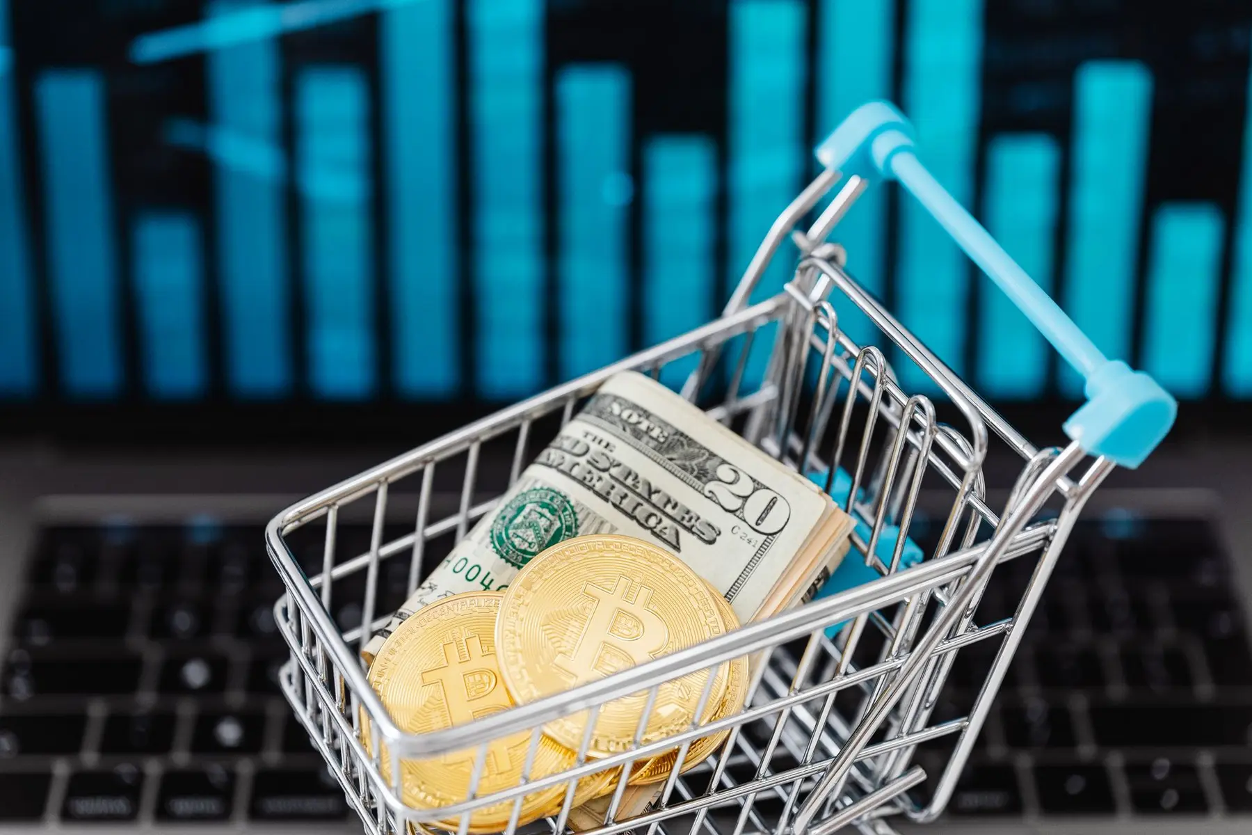 Bitcoin coins and US dollar bills inside a miniature shopping cart on a laptop keyboard with a blue chart background
