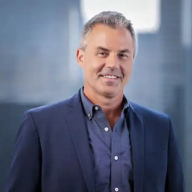Headshot of Eric Balchunas wearing a blue blazer and a button-down shirt against a blurred office background