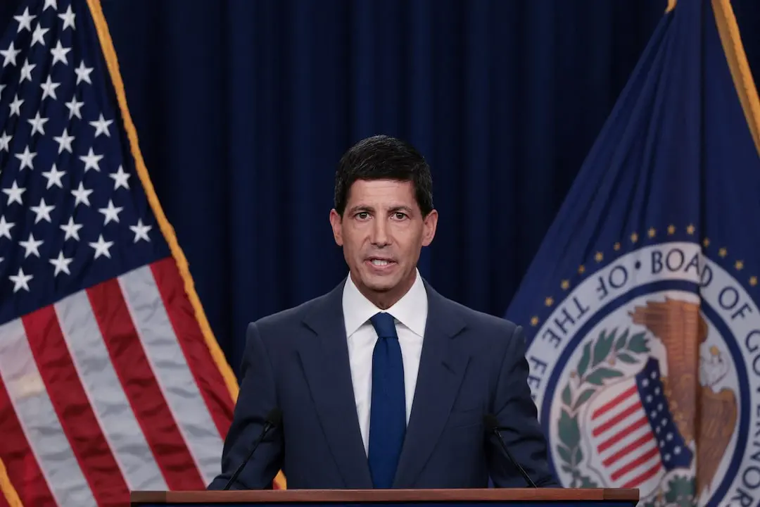 Kevin Warsh speaking at a podium with the US flag and Federal Reserve Board seal in the background