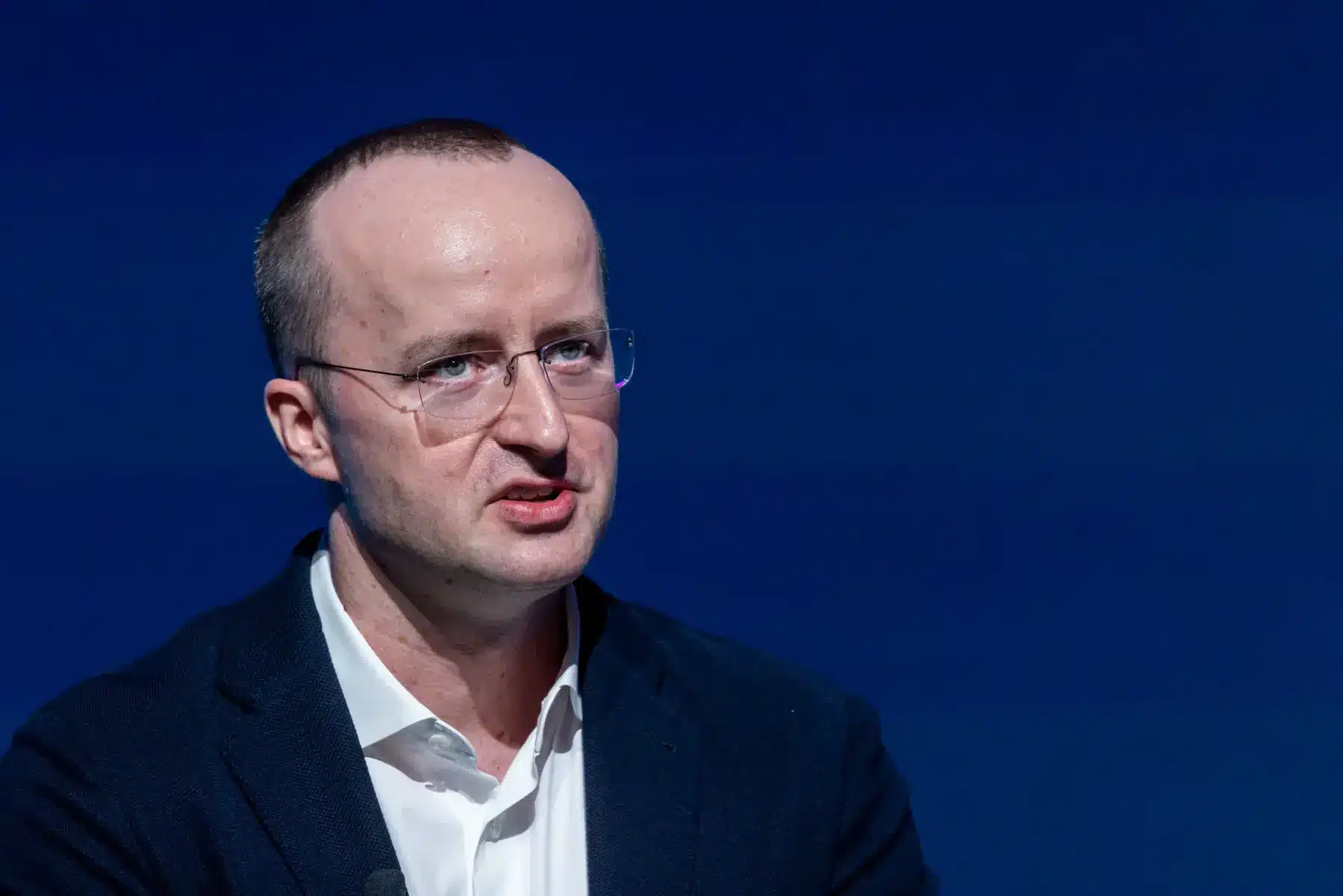 Portrait of Kris Marszalek wearing glasses and a navy blazer against a blue background