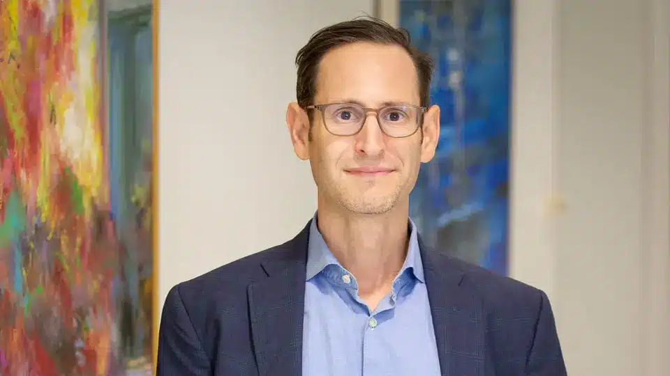 Portrait of Matthew Sigel VanEck wearing glasses and a navy blazer with a light blue shirt