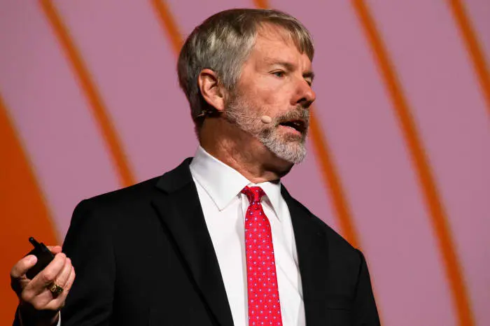 Michael Saylor speaking at a podium wearing a black suit, white shirt, and red polka-dot tie.