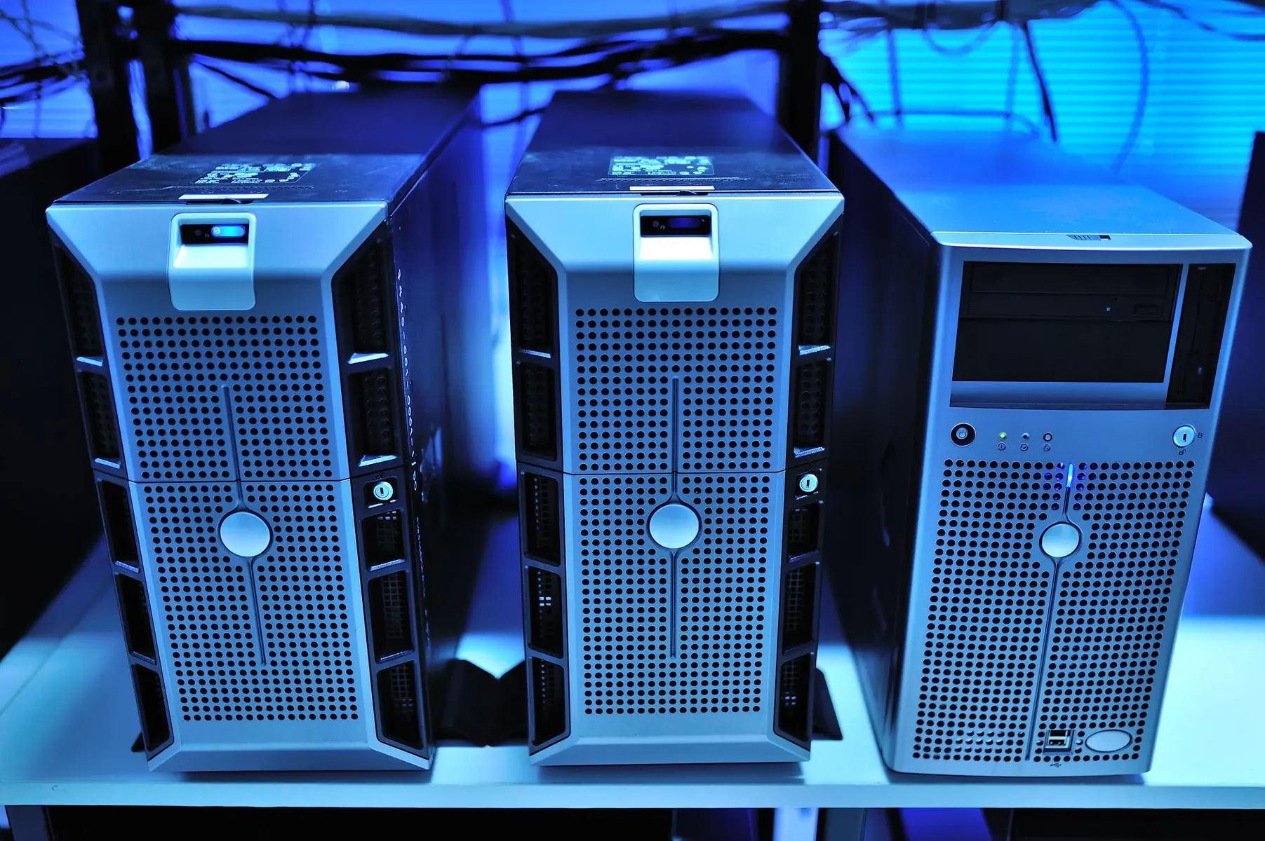 Three silver tower server computers lined up on a shelf with blue ambient lighting and cabling in the background