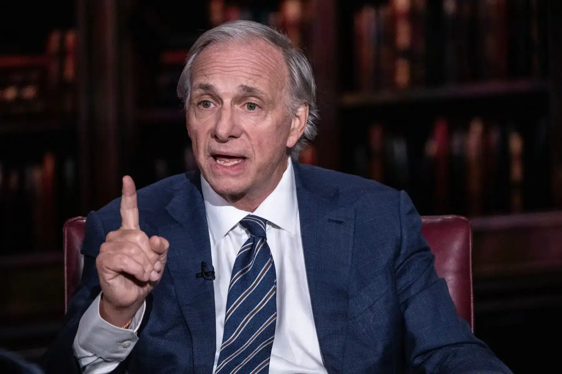 Ray Dalio wearing a blue suit and striped tie, gesturing with one finger raised during an interview