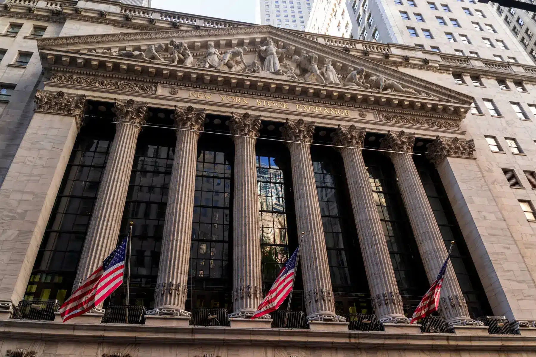 The neoclassical facade of the New York Stock Exchange building with large columns and American flags.