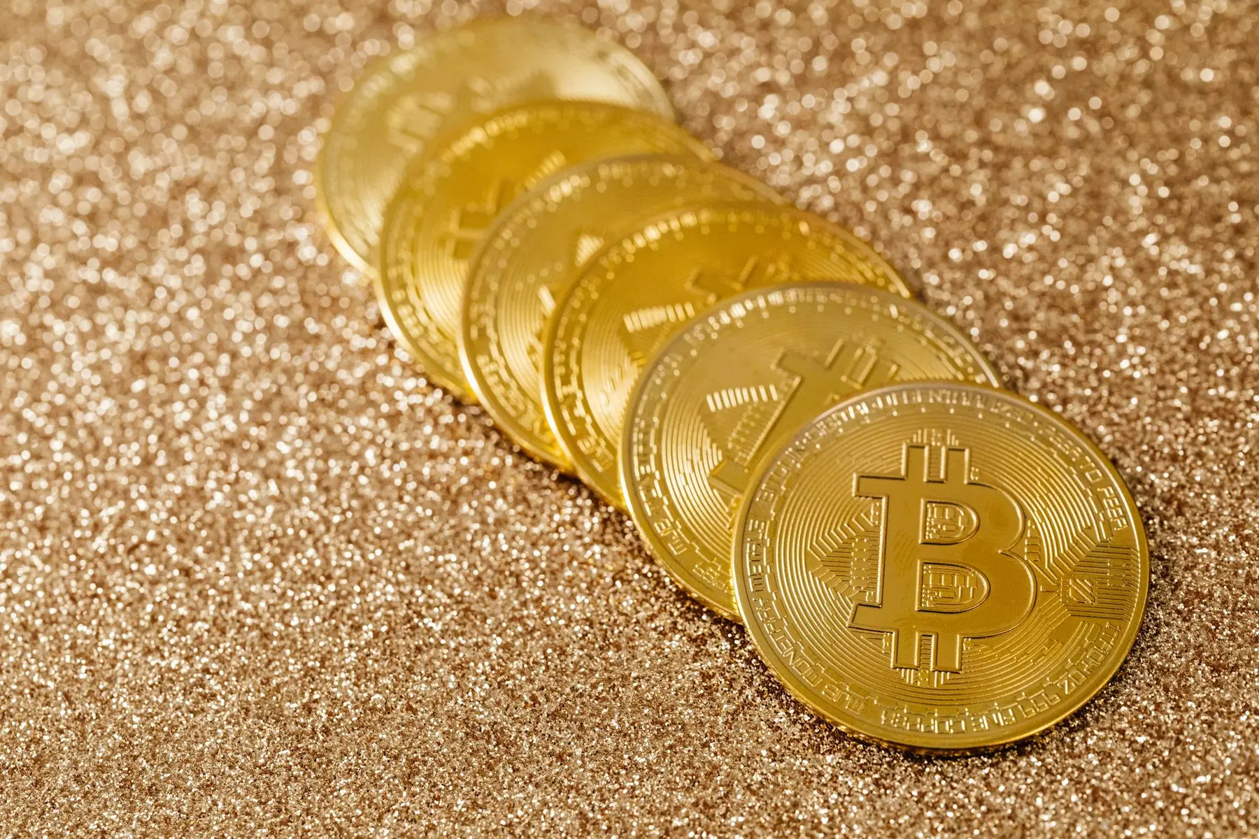 A row of gold-colored Bitcoin physical tokens resting on a shimmering gold glitter background