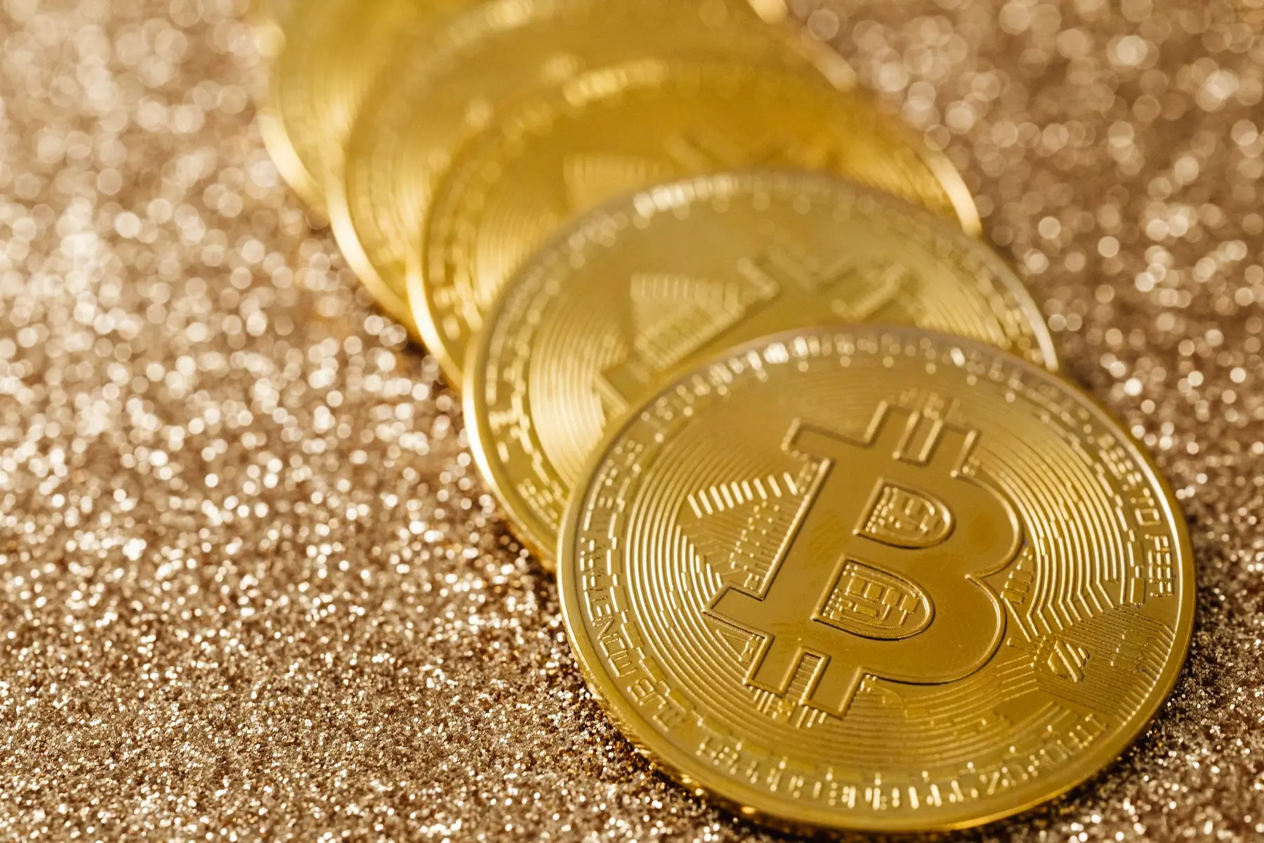 A row of gold-colored physical Bitcoin coins resting on a shimmering gold glitter surface