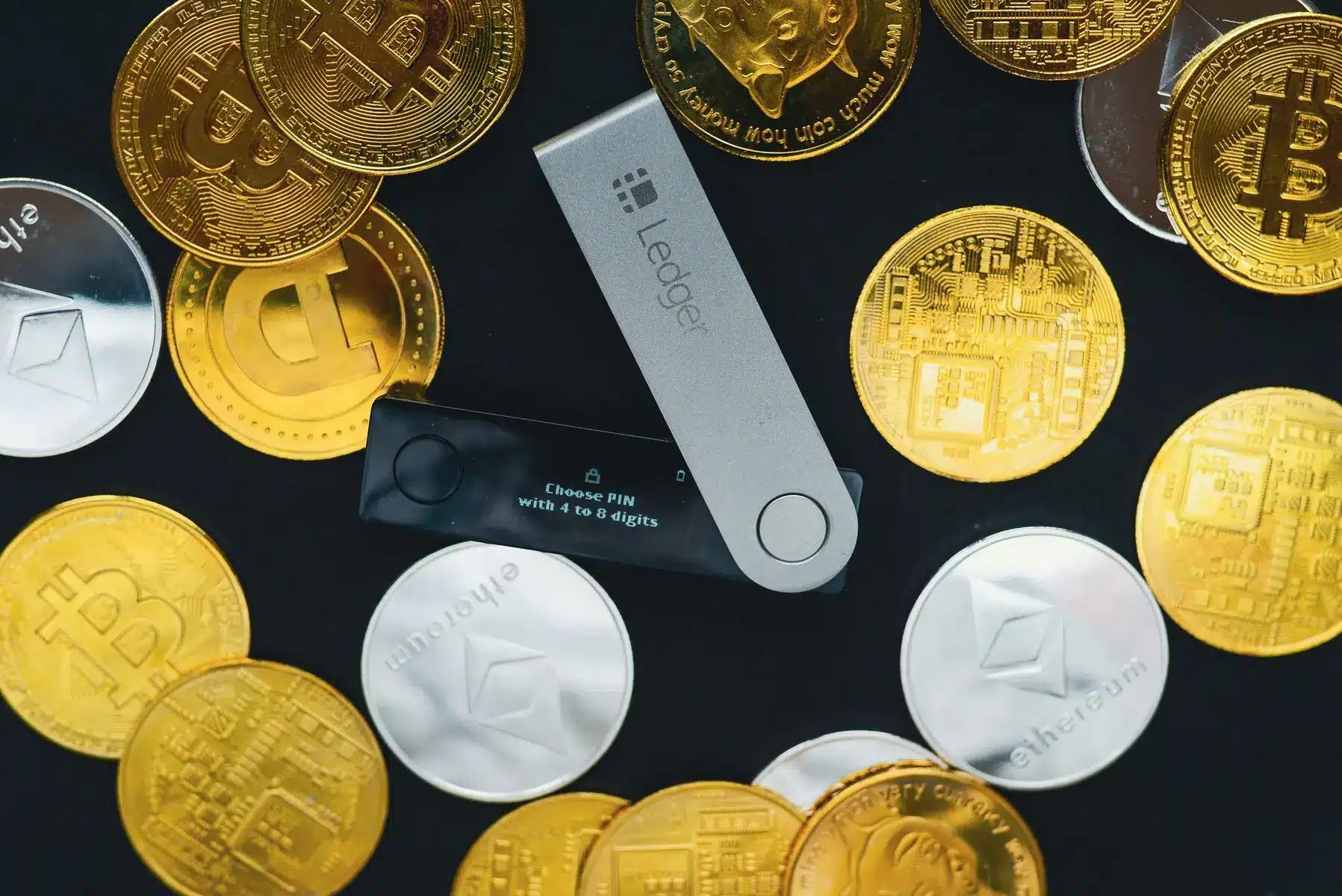 Ein Ledger Hardware-Wallet liegt auf einem schwarzen Hintergrund, umgeben von verschiedenen physischen Kryptowährungs-Münzen.