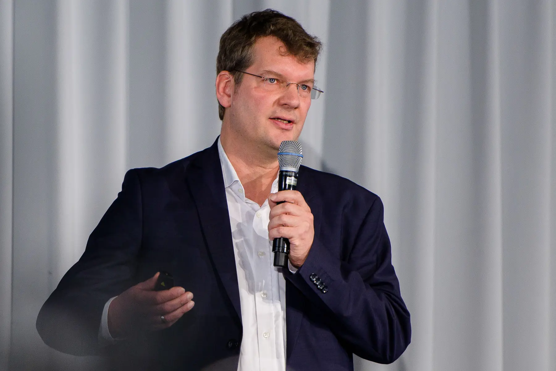 Mann mit Brille in dunkelblauem Sakko und weißem Hemd spricht in ein Mikrofon.