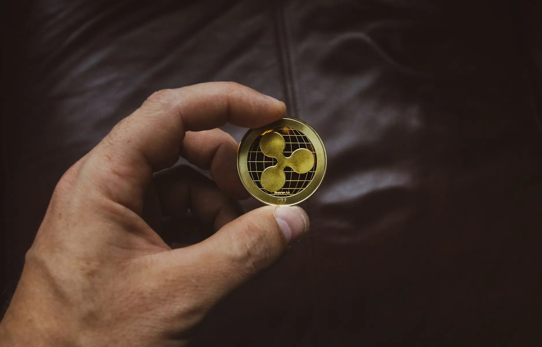 A hand holding a physical gold-colored coin featuring the Ripple XRP logo