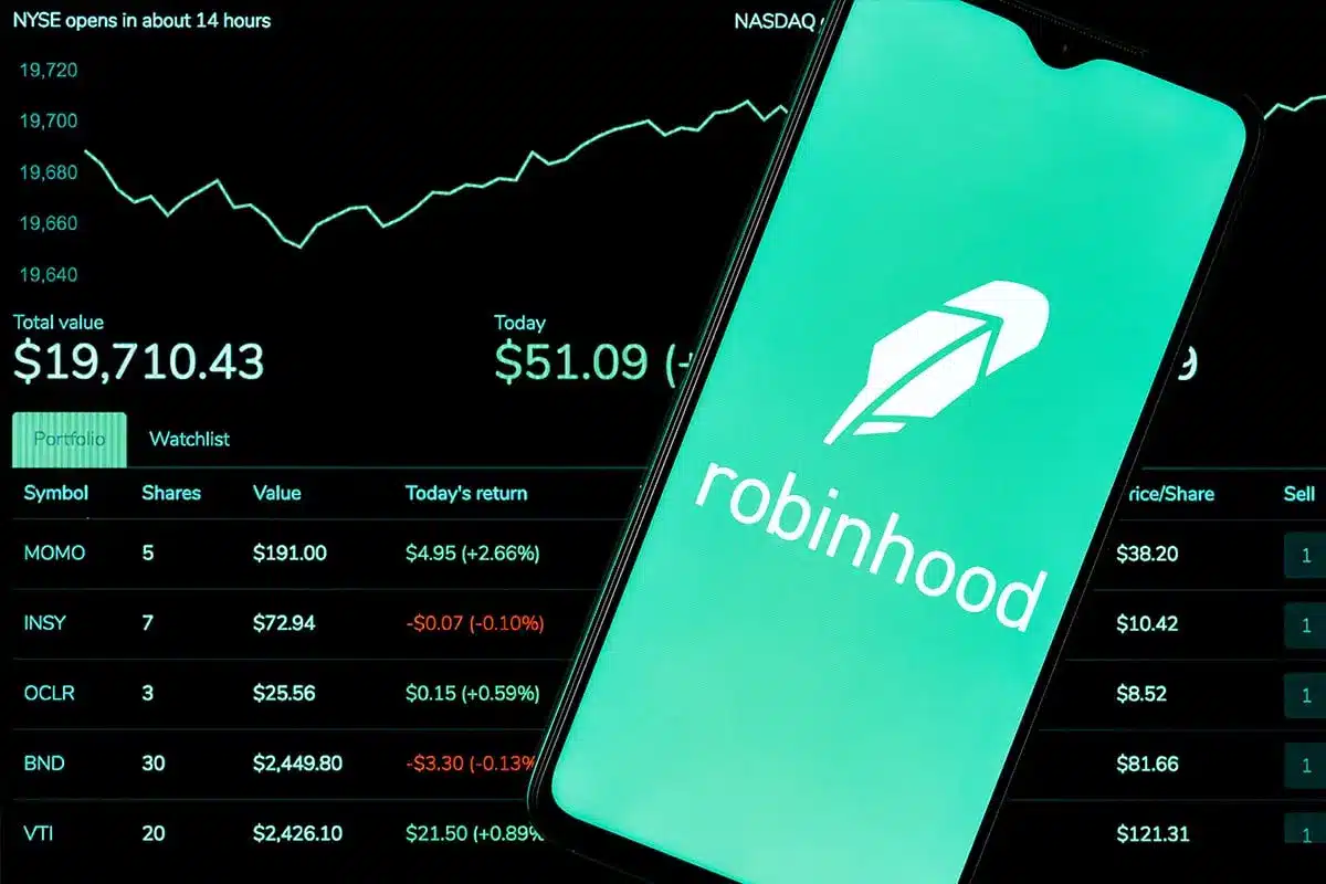 A smartphone displaying the Robinhood logo positioned over a dark background with stock market data and charts.