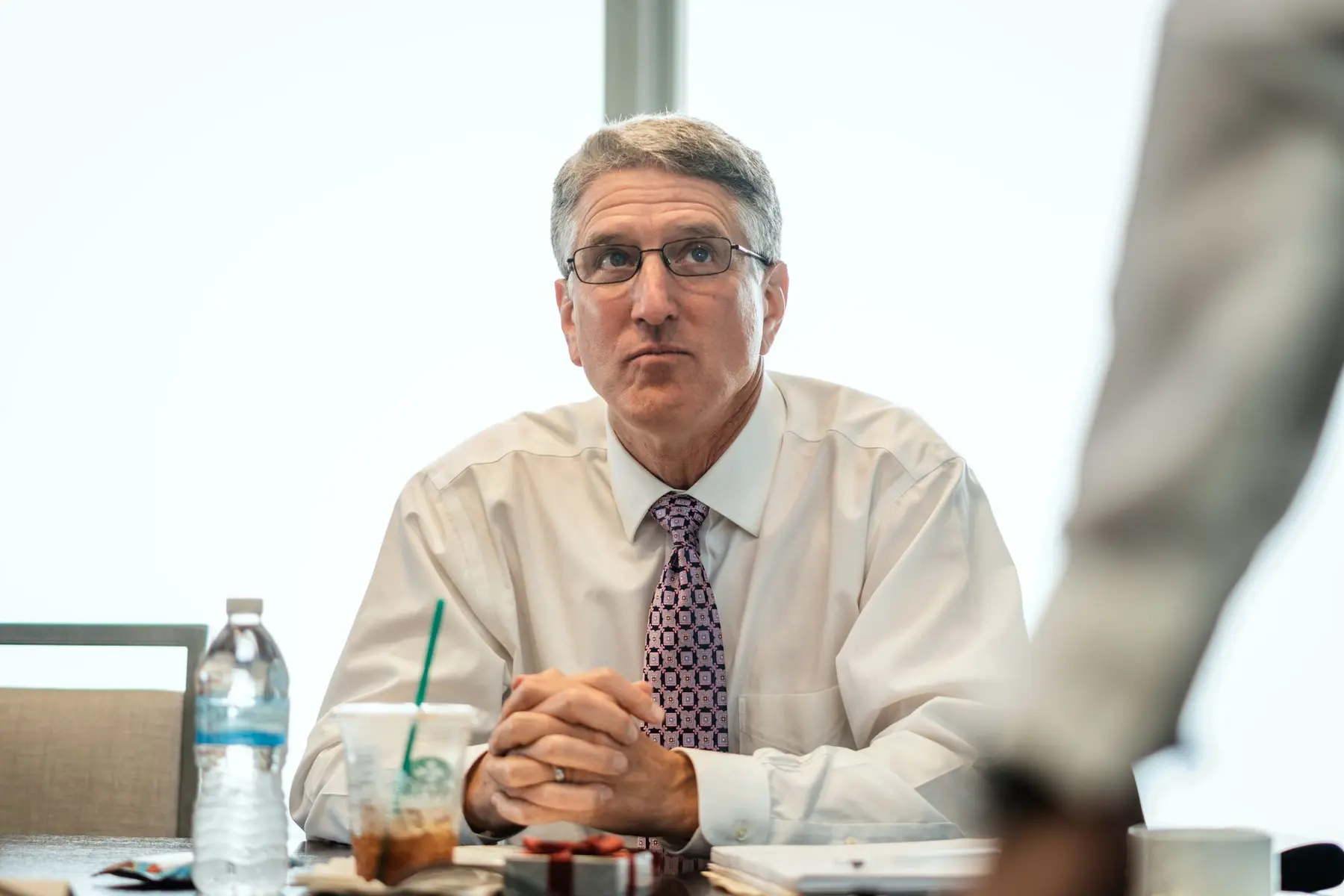 Scott Bessent wearing glasses, a white dress shirt, and a purple patterned tie, sitting at a table.