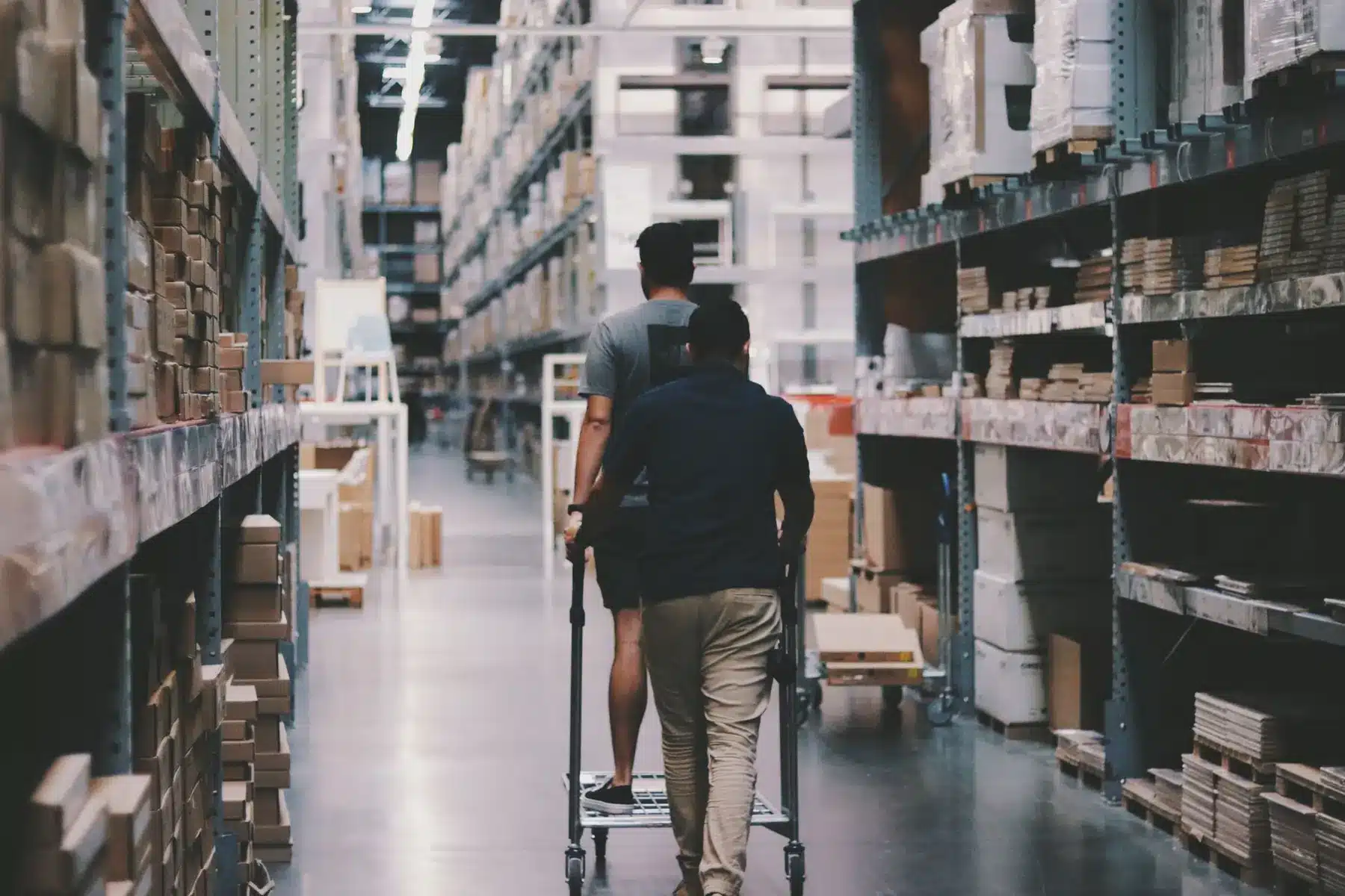 Two warehouse workers pushing a cart through an aisle of tall storage shelves filled with boxes