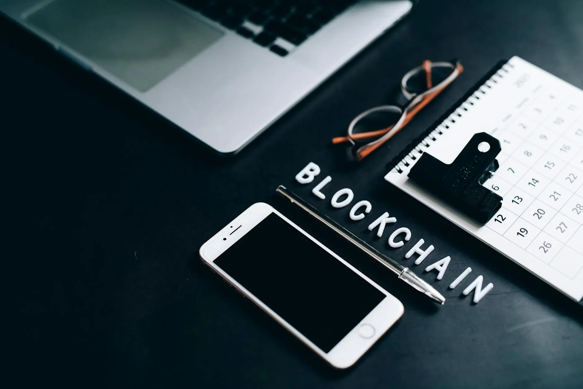 A desk with a laptop, a smartphone, glasses, and a notepad displaying 'BLOCKCHAIN'.