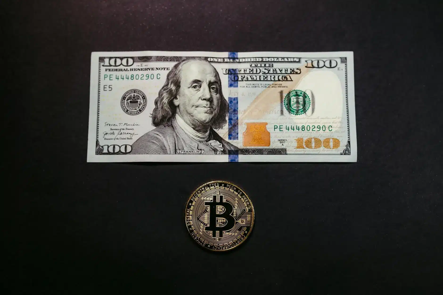A $100 bill and a Bitcoin coin placed on a dark background.
