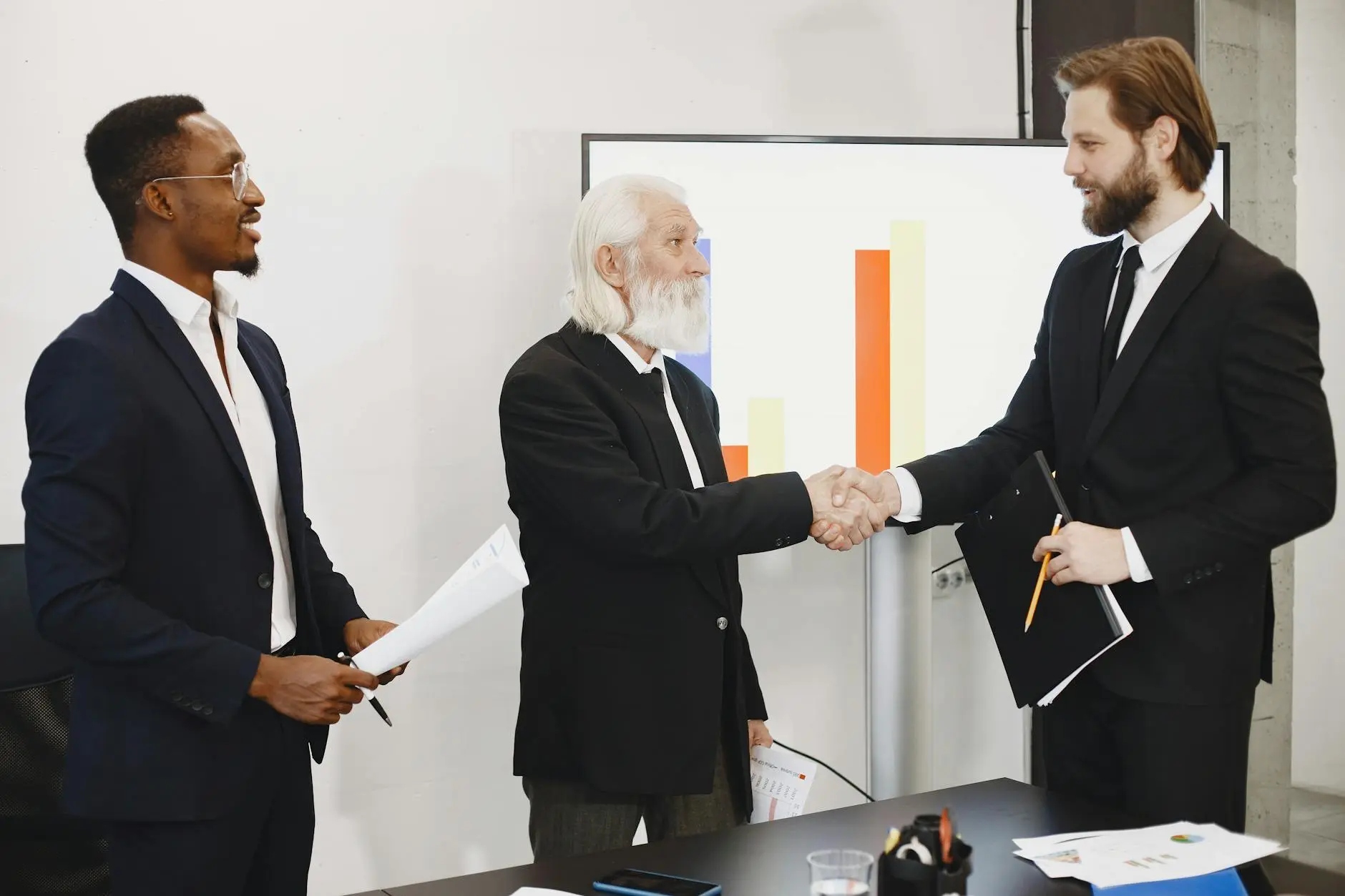 Three professionals in a meeting, two men shaking hands with a presentation screen behind them.
