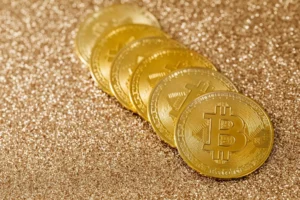 A row of gold-colored Bitcoin physical coins resting on a shimmering gold glitter surface